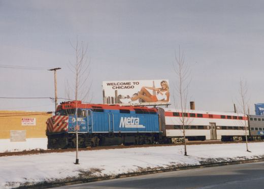 A blue and red Metra locomotive pulls a passenger train on a snow-lined track under a 'WELCOME TO CHICAGO' billboard.
