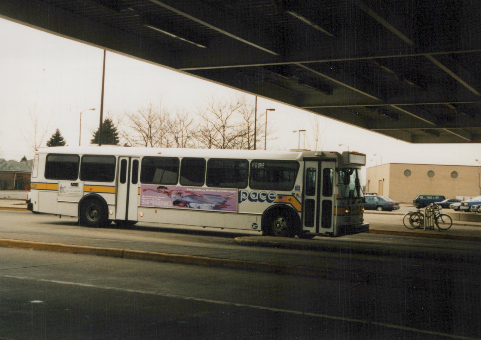 A white Pace bus, number 2326, is parked on a street under an overpass. The side of the bus features an ad for the Joffrey Ballet of Chicago. The trees in the background are bare, and a few cars and a bicycle are visible.