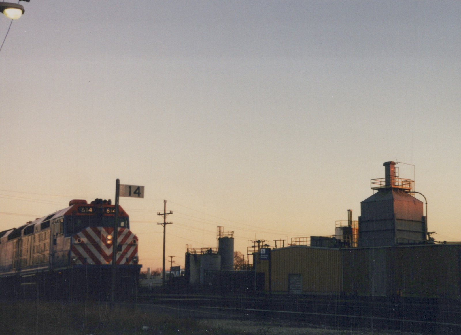 A Metra diesel locomotive numbered 614 is shown at dusk in a rail yard, with industrial buildings and a trackside sign reading '14' in the background.