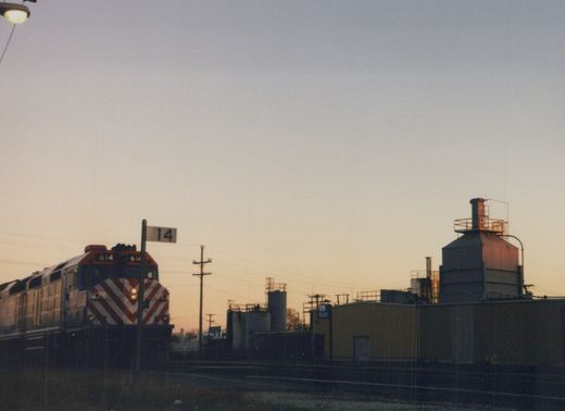A Metra diesel locomotive numbered 614 is shown at dusk in a rail yard, with industrial buildings and a trackside sign reading '14' in the background.