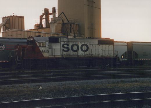 A white and maroon Soo Line diesel locomotive with the number 4413 on its side, pulling freight cars in a rail yard at dusk with industrial buildings in the background.