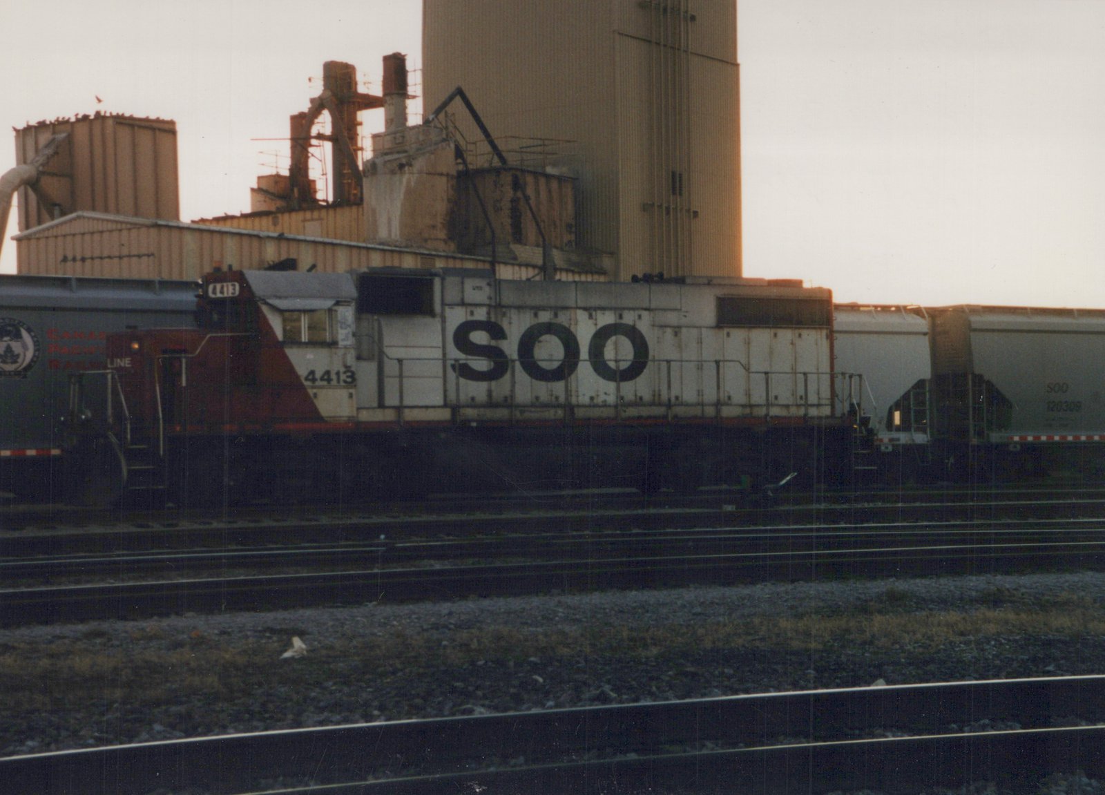 A white and maroon Soo Line diesel locomotive with the number 4413 on its side, pulling freight cars in a rail yard at dusk with industrial buildings in the background.