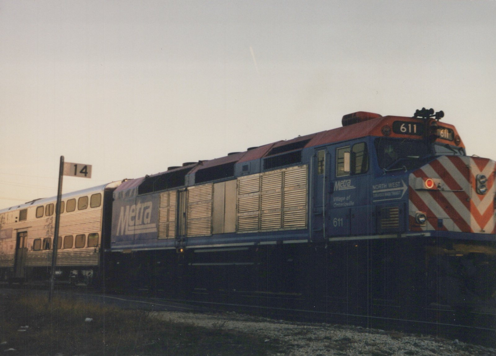 A side view of a blue and red Metra locomotive, number 611, pulling silver commuter cars on a railroad track under a cloudy sky.