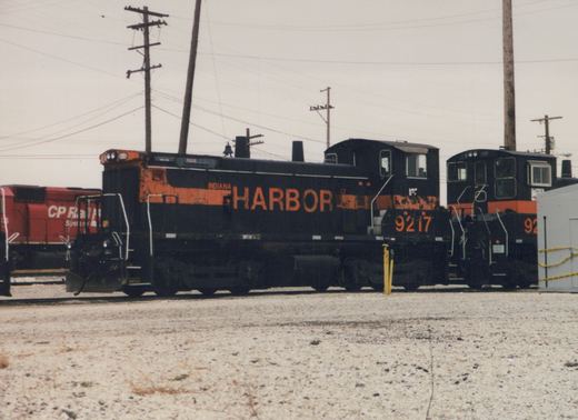 Side view of two black and orange Indiana Harbor Belt Railroad locomotives, number 9217 clearly visible, parked in a gravel yard with a red CP Rail locomotive in the background under an overcast sky.