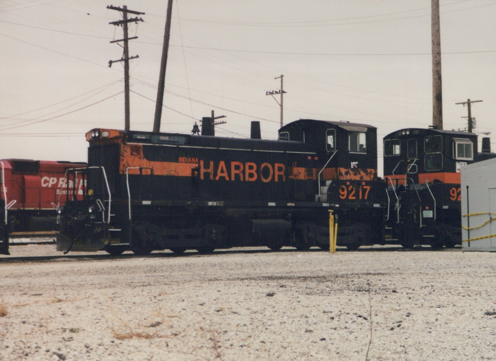 Side view of two black and orange Indiana Harbor Belt Railroad locomotives, number 9217 clearly visible, parked in a gravel yard with a red CP Rail locomotive in the background under an overcast sky.