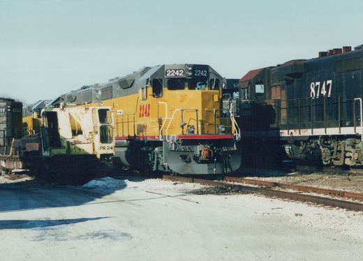 A yellow locomotive and a black Illinois Central locomotive numbered 8747 sit on parallel tracks in a snowy yard, with a white maintenance vehicle nearby.