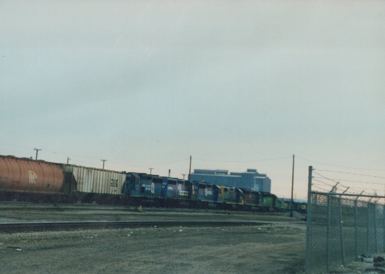 A long freight train with CSX locomotives and freight cars running through a fenced yard at dusk.