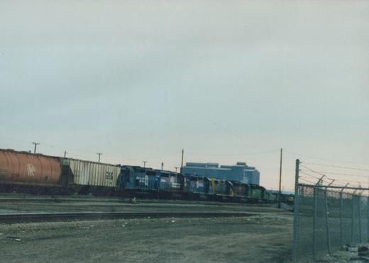 A long freight train with CSX locomotives and freight cars running through a fenced yard at dusk.