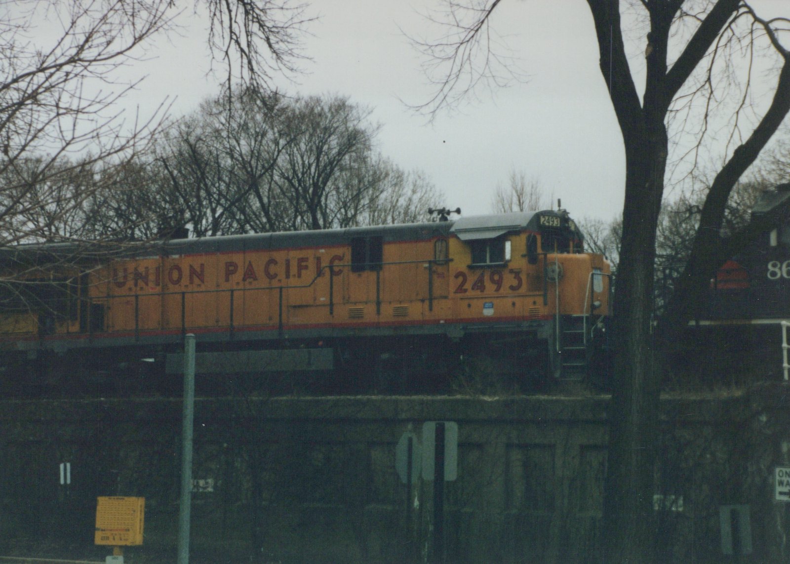 A yellow Union Pacific locomotive with number 2493 on its side and front is traveling along an elevated track under an overcast sky, with bare trees in the background and a partial view of another locomotive behind it.