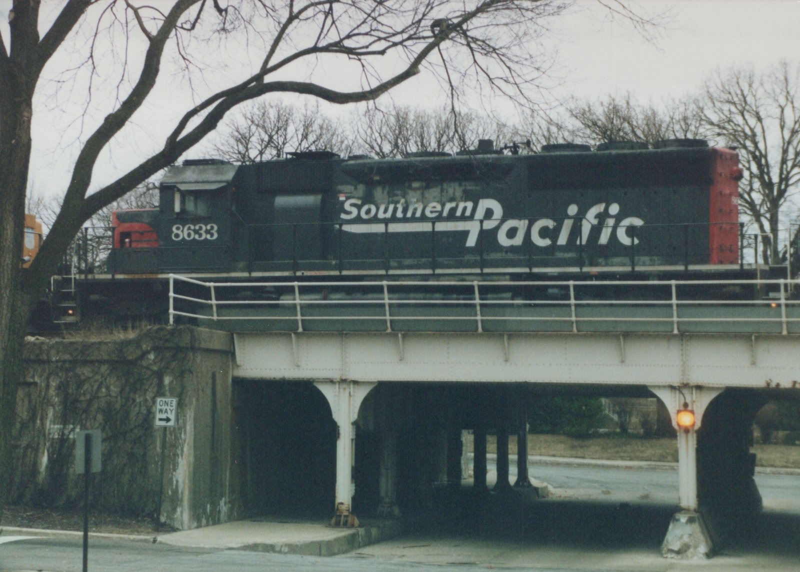 A black Southern Pacific diesel locomotive numbered 8633 travels on an elevated steel bridge; below, a street sign reads ONE WAY and bare trees frame the scene.