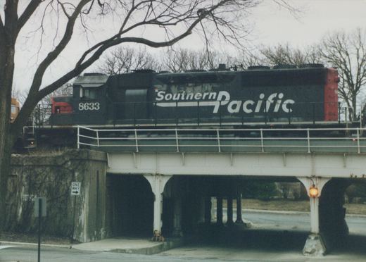 A black Southern Pacific diesel locomotive numbered 8633 travels on an elevated steel bridge; below, a street sign reads ONE WAY and bare trees frame the scene.