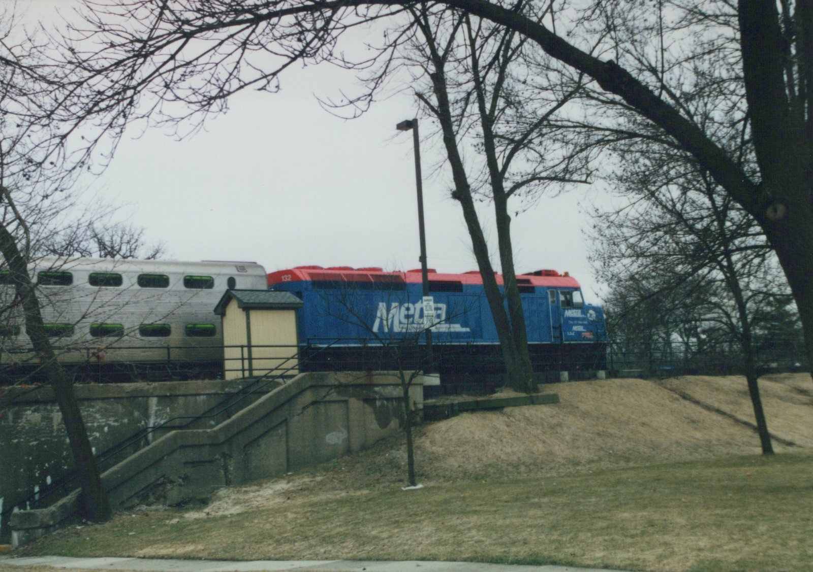 A blue and red Metra locomotive numbered 132 is visible on a raised track bed with bare trees and a concrete staircase nearby on an overcast day.
