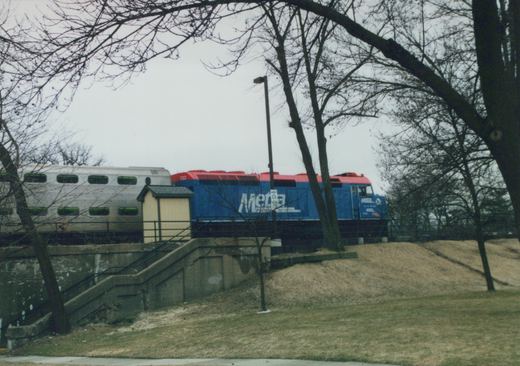 A blue and red Metra locomotive numbered 132 is visible on a raised track bed with bare trees and a concrete staircase nearby on an overcast day.