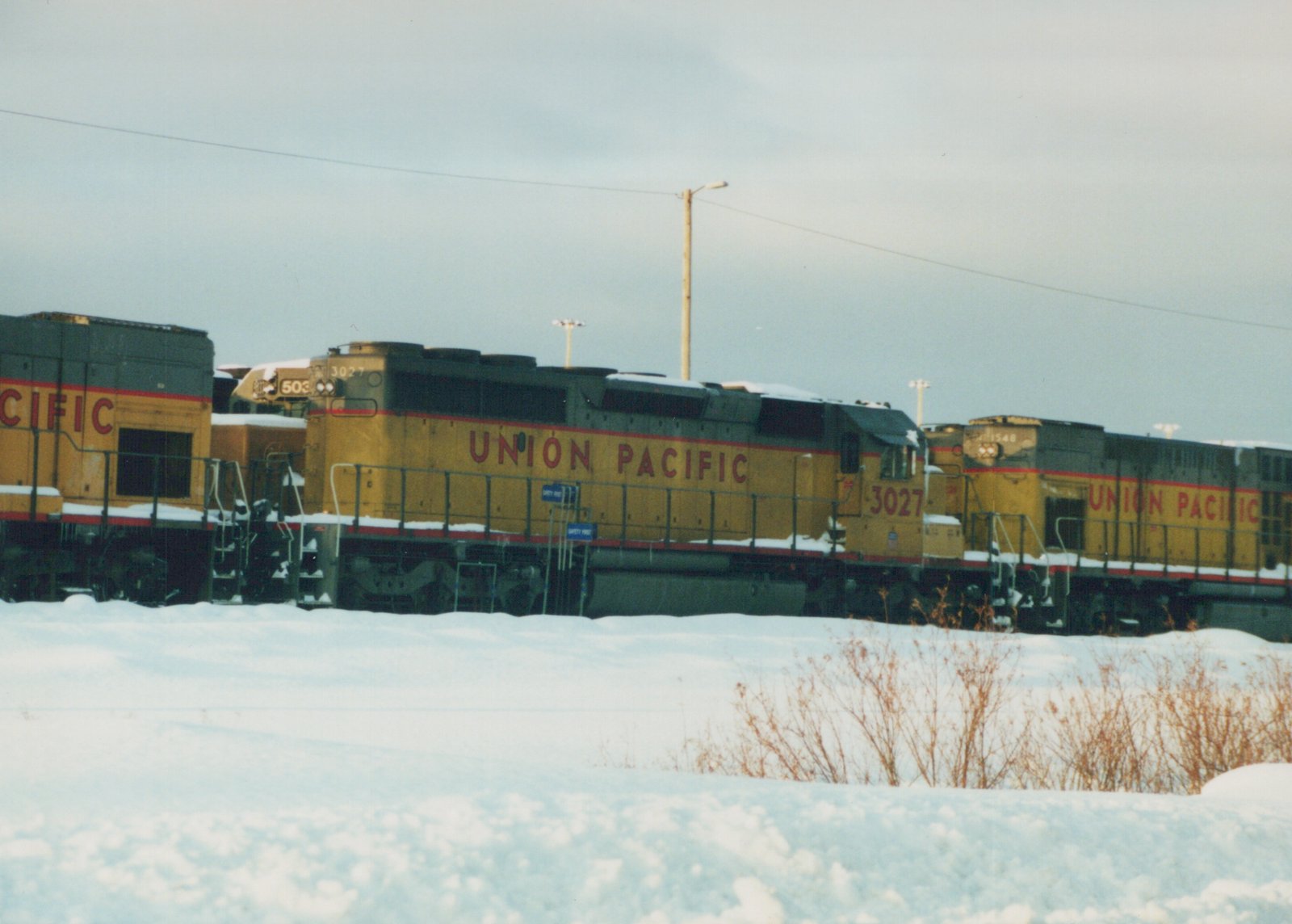 A side view of three yellow and gray Union Pacific diesel locomotives, numbered 3027, 503, and 1548, parked on a snow-covered track with a cloudy sky.