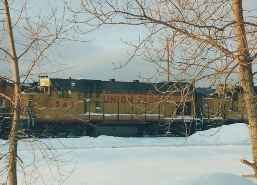 A yellow and black Union Pacific diesel locomotive numbered 542 sits on a snow-covered track, partially framed by bare tree branches.