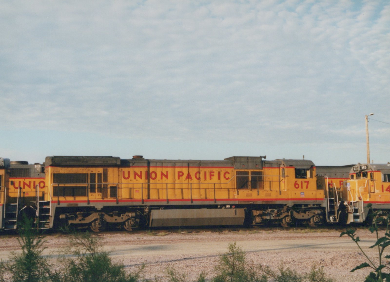Side view of two yellow Union Pacific diesel locomotives, numbers 617 and 143, parked in a gravel yard with a cloudy sky above.