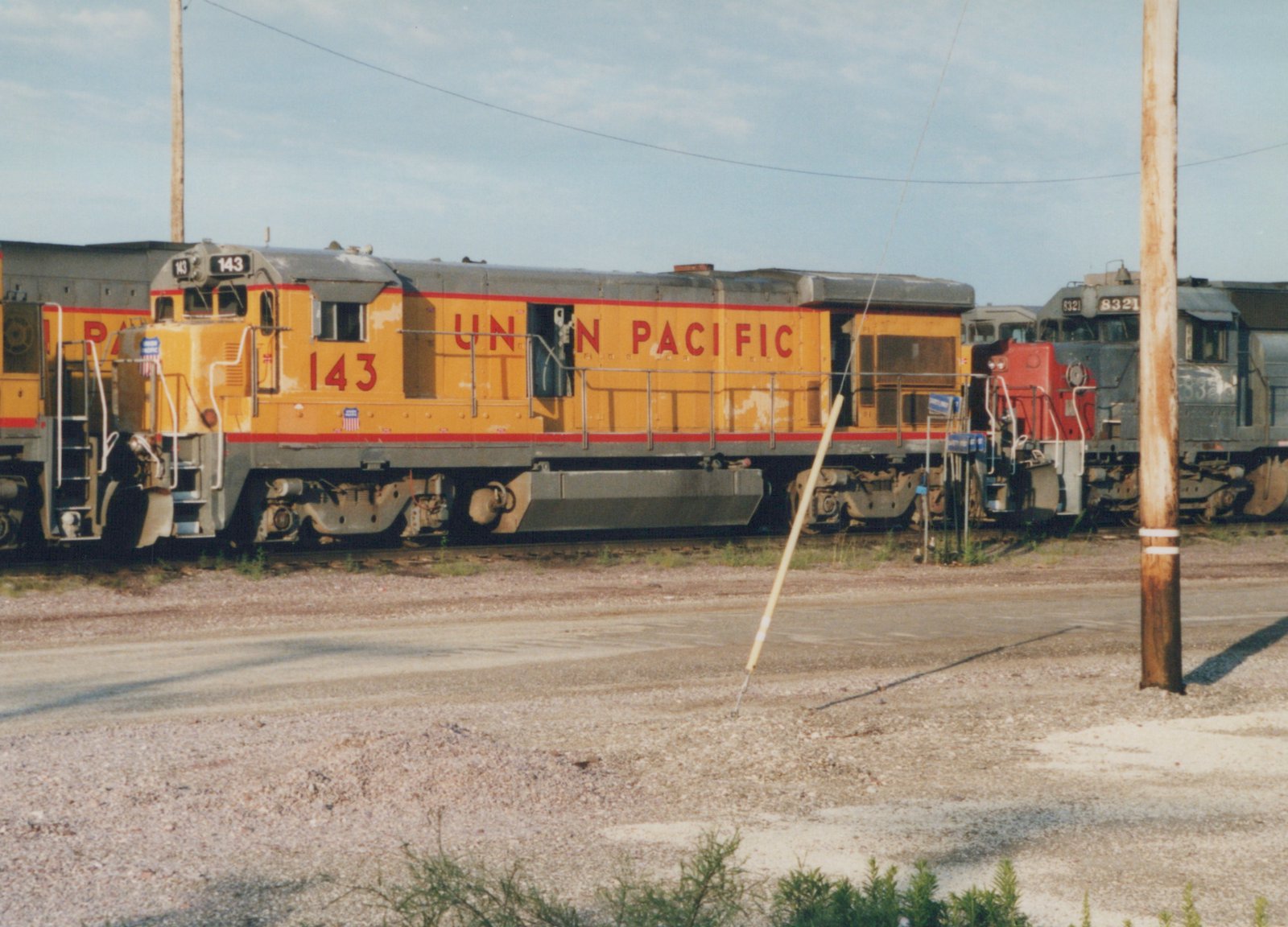 Side view of a yellow Union Pacific locomotive numbered 143 parked in a gravel yard with another gray locomotive behind it under a clear sky.