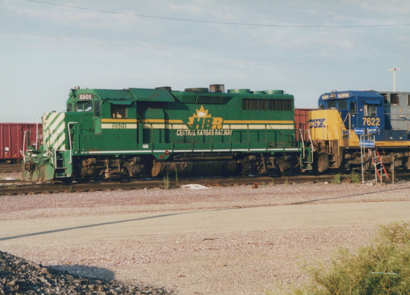 A green diesel locomotive with the number 2501 and Central Kansas Railway lettering is parked on tracks next to a blue and yellow CSX locomotive in a gravel yard under a clear sky.