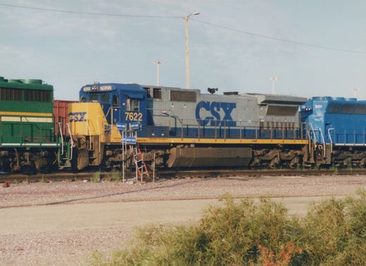A side view of a blue and gray CSX diesel locomotive with the number 7622, coupled to another locomotive and a freight car in a gravel yard.