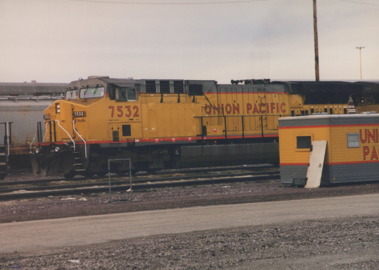 A yellow Union Pacific diesel locomotive numbered 7532 is parked on tracks in a gravel yard, with a matching trailer nearby and other freight cars in the background under a gray sky.