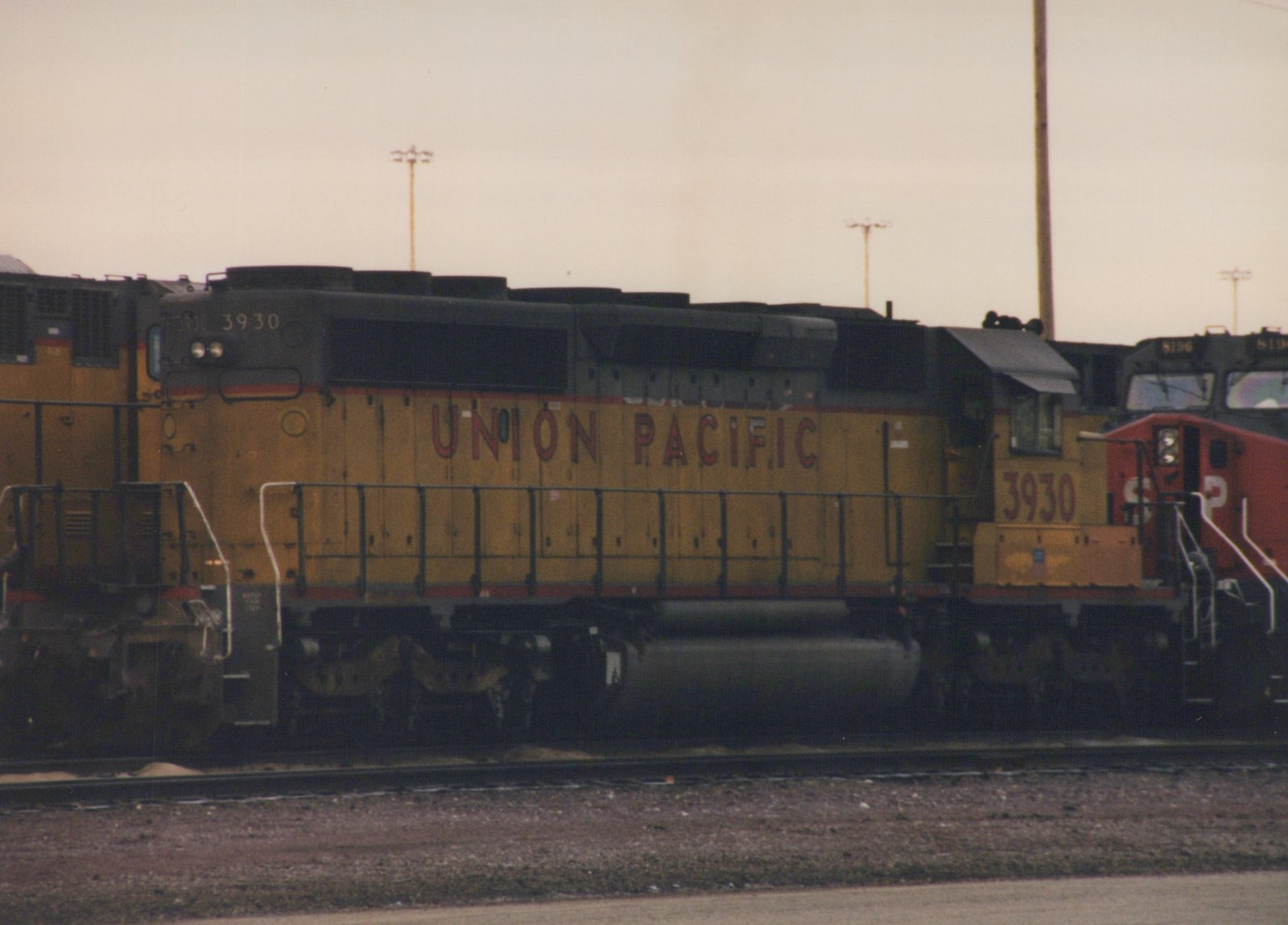A side view of a Union Pacific diesel locomotive, number 3930, painted in yellow with a red stripe, parked in a rail yard. A red and black Southern Pacific locomotive is visible in the background.