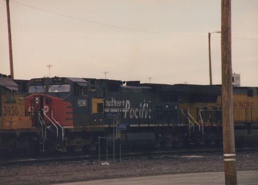 A black and red Southern Pacific locomotive, number 8196, is parked on tracks at a rail yard with other locomotives behind it.