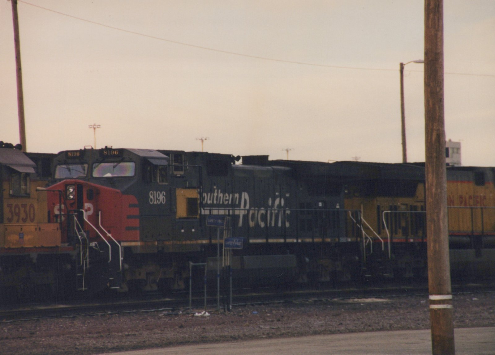 A black and red Southern Pacific locomotive, number 8196, is parked on tracks at a rail yard with other locomotives behind it.