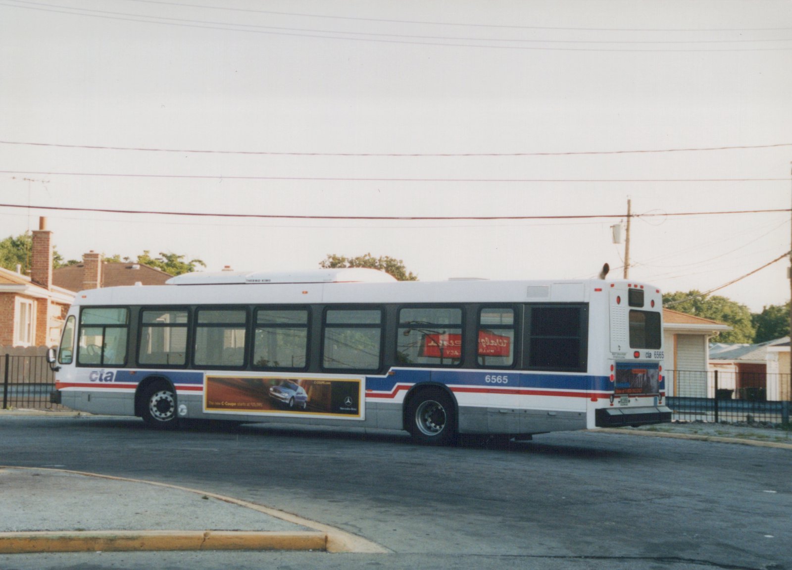 A white, blue, and red Chicago Transit Authority bus, number 6565, is parked on an asphalt street in front of residential houses and a fence.