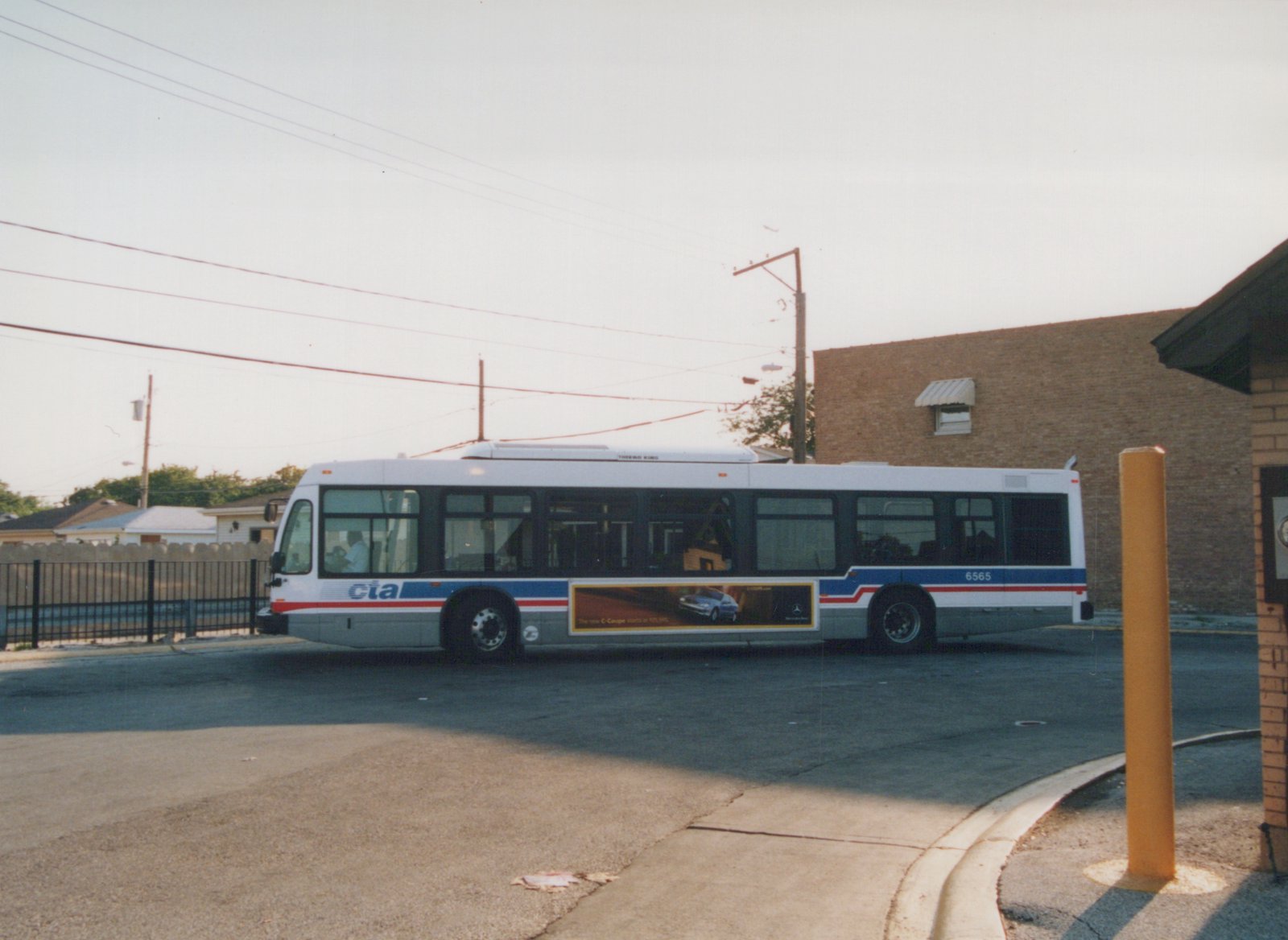 Side view of a white Chicago Transit Authority bus with a blue and red stripe, displaying the number 6565 and a Mercedes-Benz advertisement, parked on a paved street next to a brick building.