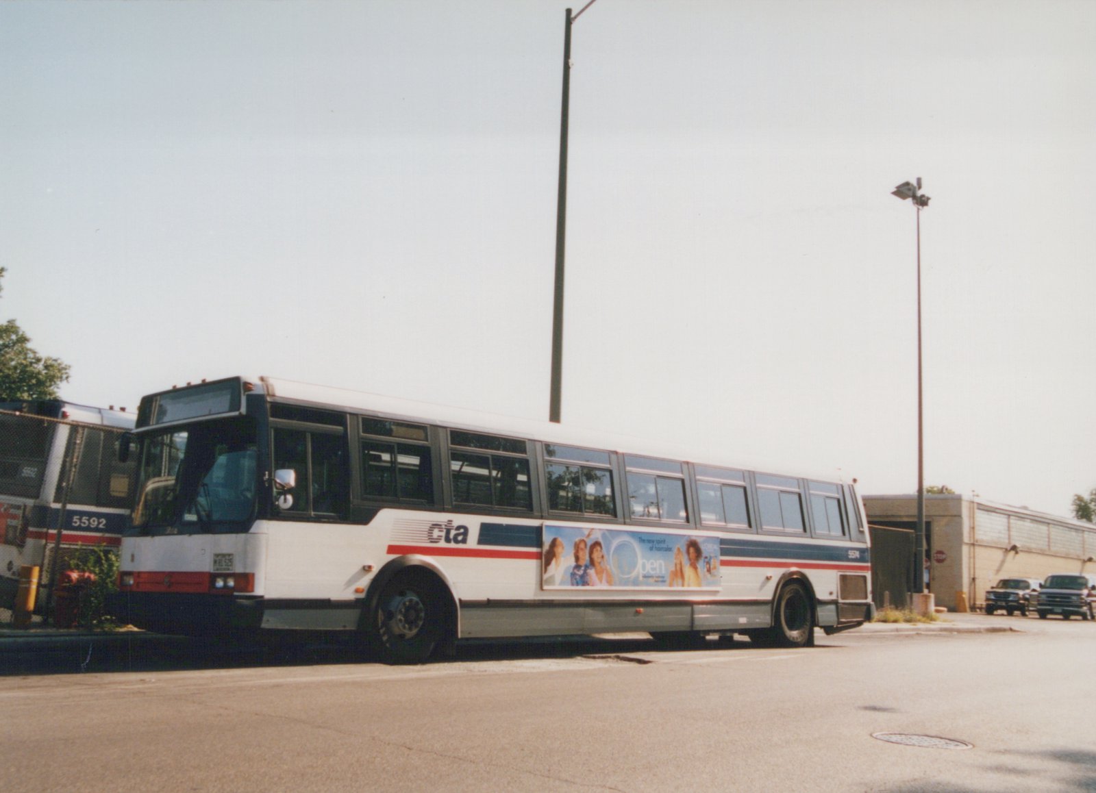 Side view of a white Chicago Transit Authority bus with red and blue stripes, number 5574, parked in a lot with another bus and trucks nearby.