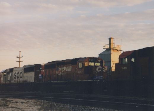 Three diesel locomotives, a Soo Line 6017, a CP Rail 5667, and a CP Rail 748, are coupled together in a railroad yard at dusk with an industrial building in the background.