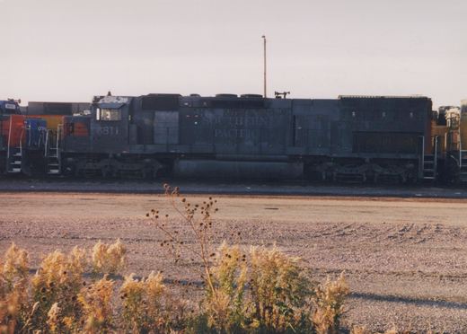 A side view of a dark gray Southern Pacific diesel locomotive, number 6811, parked in a gravel yard with dry autumn weeds in the foreground.