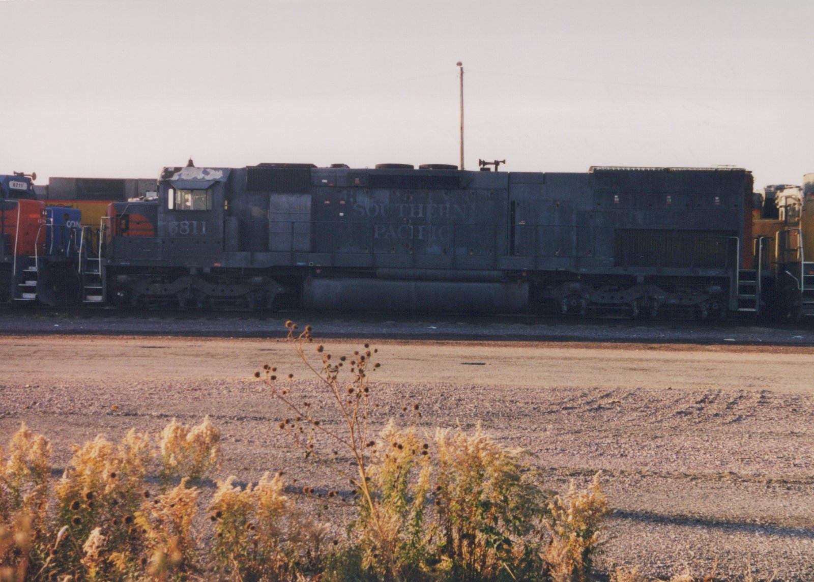 A side view of a dark gray Southern Pacific diesel locomotive, number 6811, parked in a gravel yard with dry autumn weeds in the foreground.