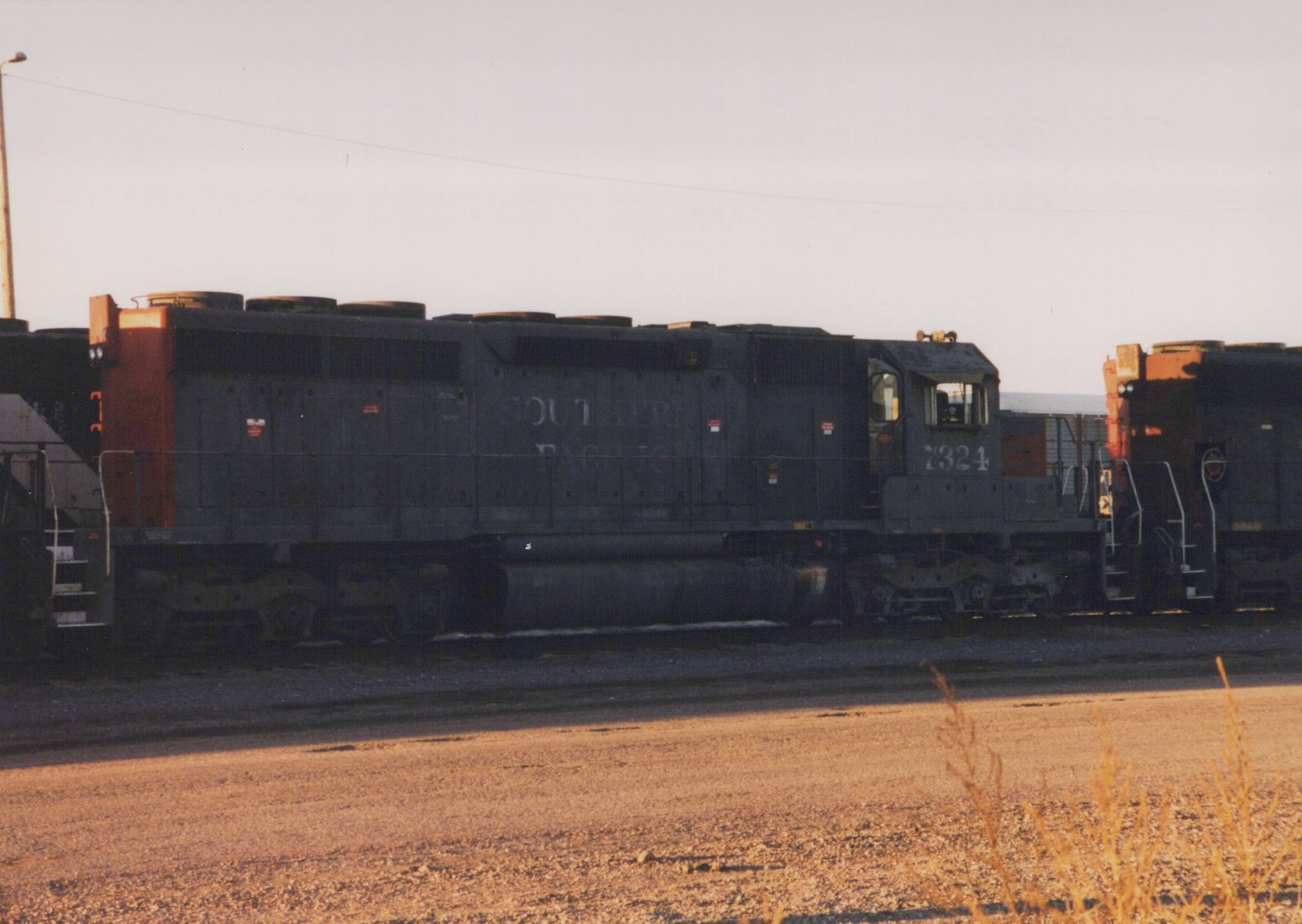 A black and red Southern Pacific diesel locomotive, number 7324, is parked on a track in a gravel yard at dusk, with another locomotive coupled behind it.