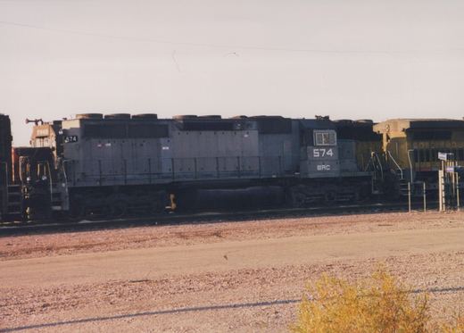 A gray diesel locomotive numbered 574 with BRC lettering is parked on a track in a gravel yard on a clear day, with other locomotives visible in the background.