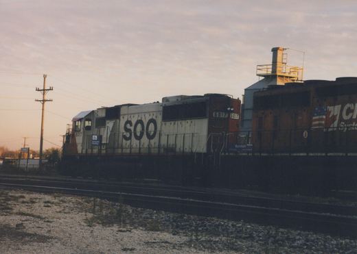 A white SOO Line locomotive with the number 6017 on the side is stopped at a station platform, with a dark Canadian National locomotive next to it. The sky is overcast, and the ground is gravel.