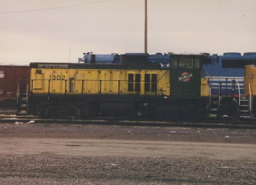 Side view of a yellow and green Chicago and North Western locomotive number 1302 in a gravel rail yard, with a red freight car and a blue and white locomotive in the background.