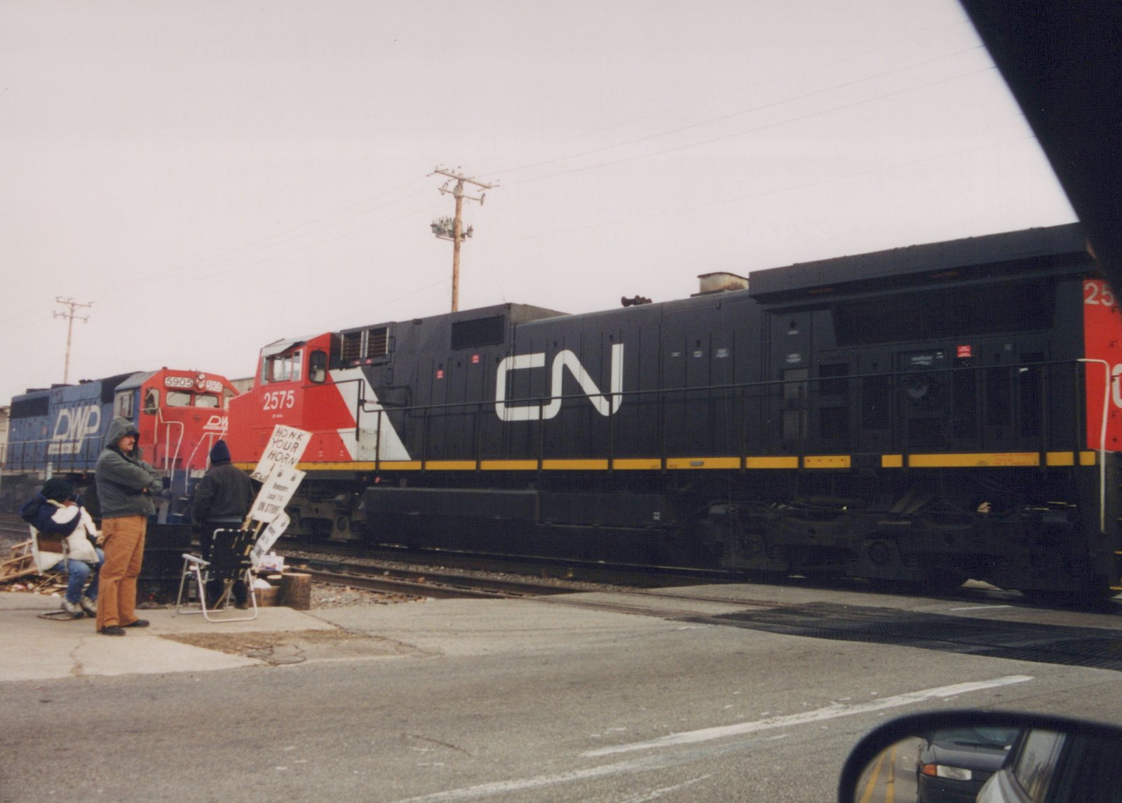 A black and yellow Canadian National GP38-2 locomotive numbered 2575 sits on the tracks at a street crossing, with a blue DWP locomotive numbered 5905 behind it, while people stand nearby holding a handwritten 'HONK YOUR HORN' sign.