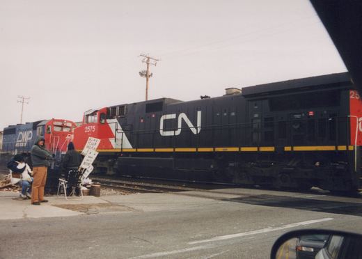 A black and yellow Canadian National GP38-2 locomotive numbered 2575 sits on the tracks at a street crossing, with a blue DWP locomotive numbered 5905 behind it, while people stand nearby holding a handwritten 'HONK YOUR HORN' sign.