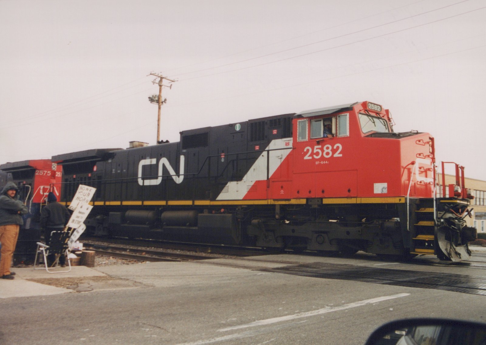 A red, black, and white Canadian National diesel locomotive numbered 2582 is stopped at a grade crossing with people standing nearby holding a sign that says 'HONK YOUR HORN.'