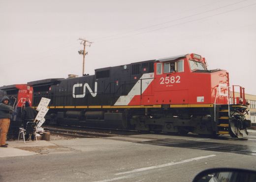 A red, black, and white Canadian National diesel locomotive numbered 2582 is stopped at a grade crossing with people standing nearby holding a sign that says 'HONK YOUR HORN.'