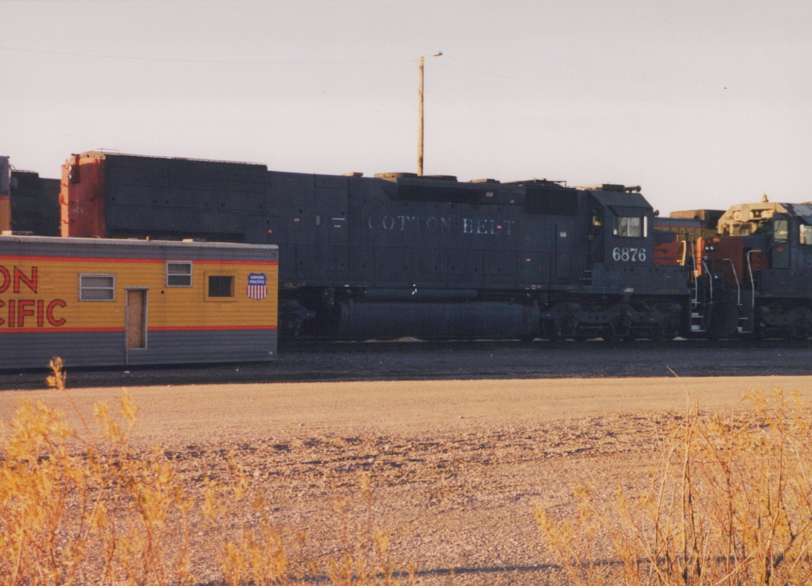 Side view of a black Cotton Belt diesel locomotive with the number 6876, parked on tracks next to a yellow Union Pacific office car in a gravel yard under a clear sky.
