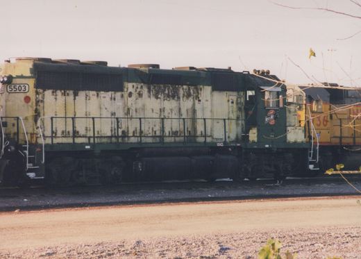 Side view of a green and yellow Chicago and North Western diesel locomotive numbered 5503, with another yellow locomotive numbered 736 behind it, parked in a gravel yard with bare tree branches in the foreground.