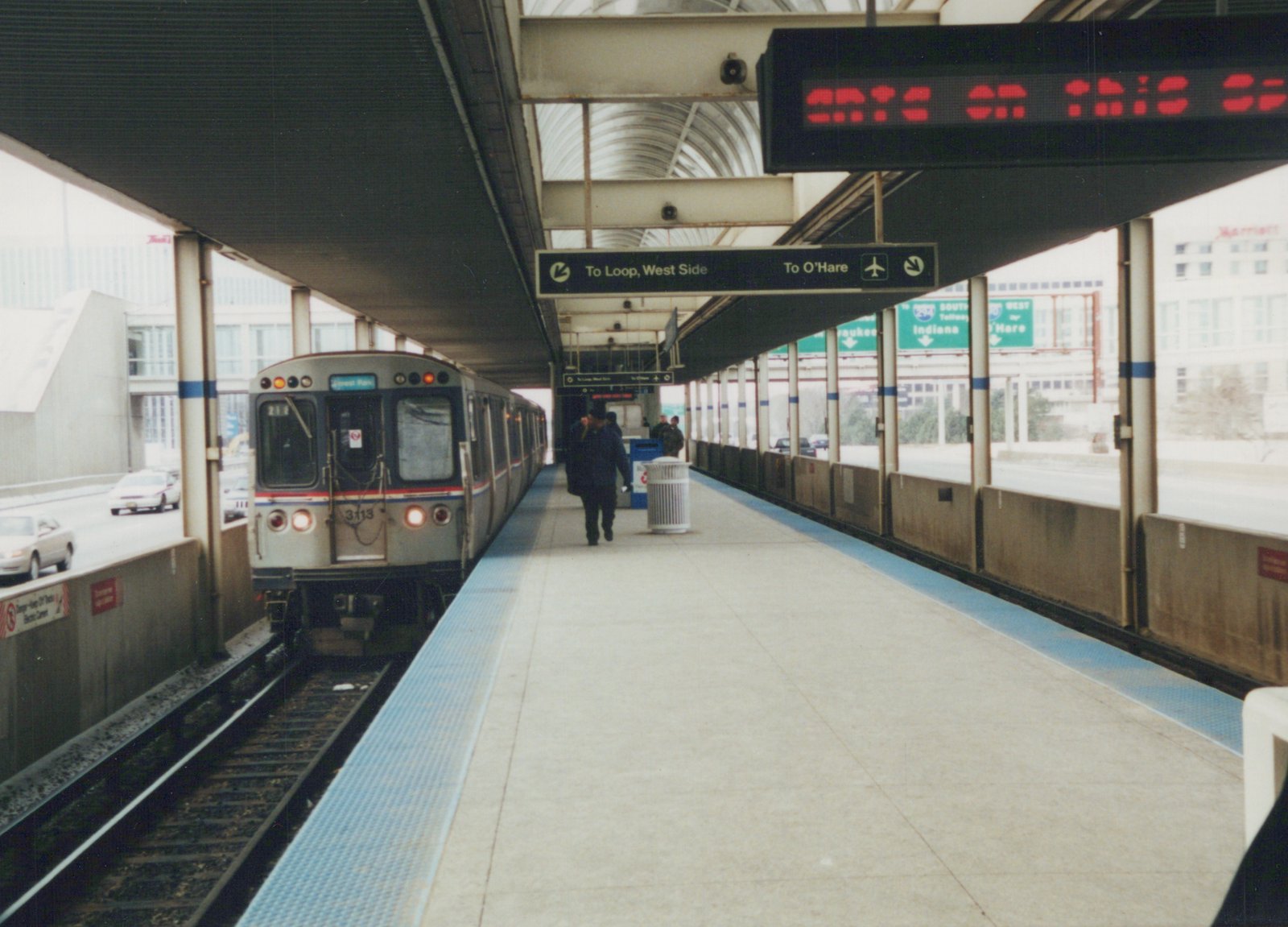 A silver Chicago Transit Authority rapid transit train, number 3113, is stopped at an elevated station platform with passengers walking nearby; highway signs for O'Hare Airport and the West Side are visible overhead.