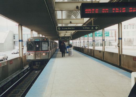 A silver Chicago Transit Authority rapid transit train, number 3113, is stopped at an elevated station platform with passengers walking nearby; highway signs for O'Hare Airport and the West Side are visible overhead.