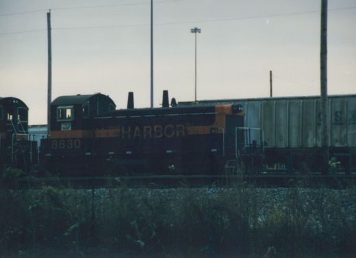 Side view of a dark green and orange Indiana Harbor Belt Railroad diesel locomotive, number 8830, in a railroad yard at dusk next to a grey freight car.