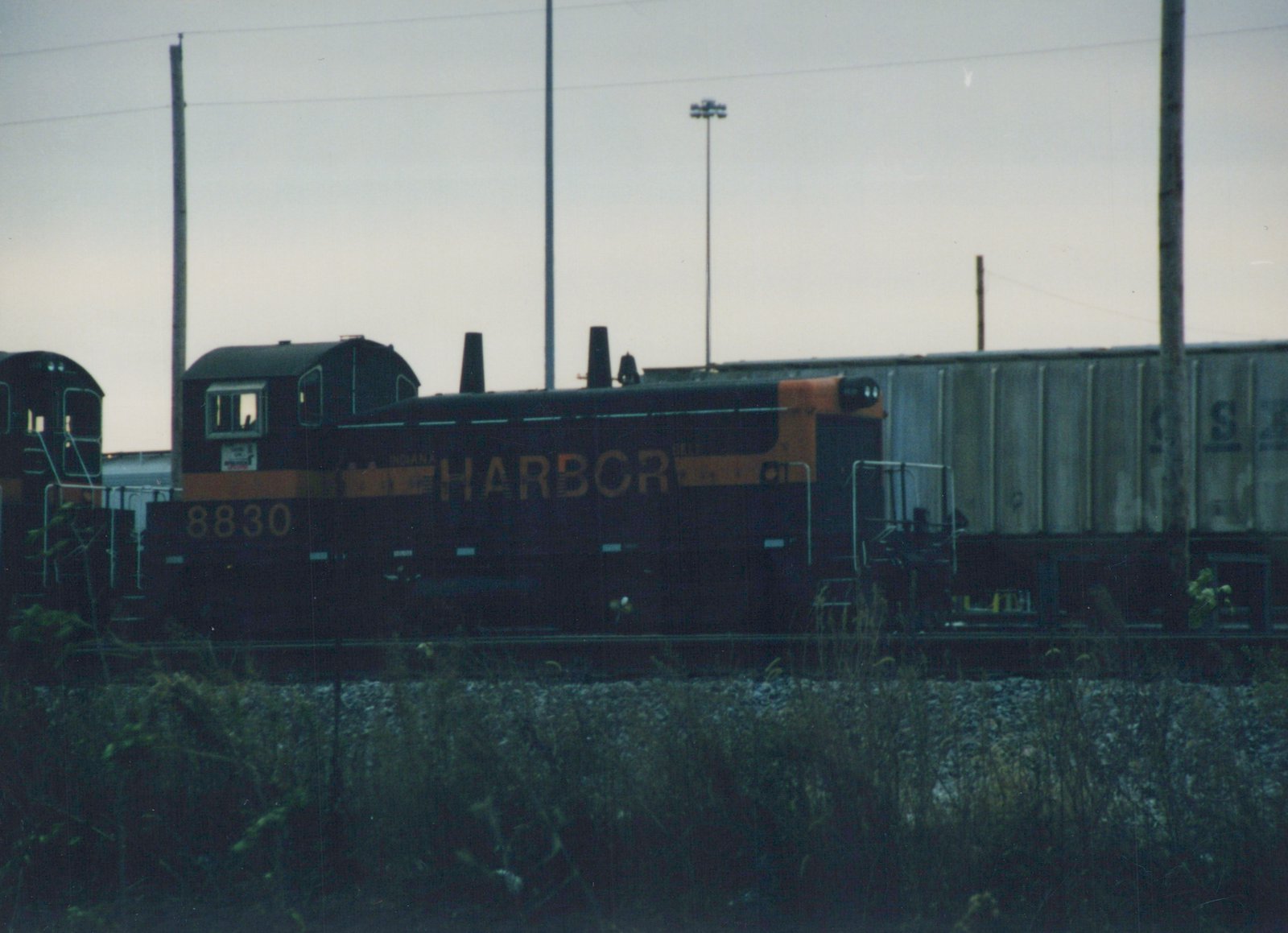 Side view of a dark green and orange Indiana Harbor Belt Railroad diesel locomotive, number 8830, in a railroad yard at dusk next to a grey freight car.