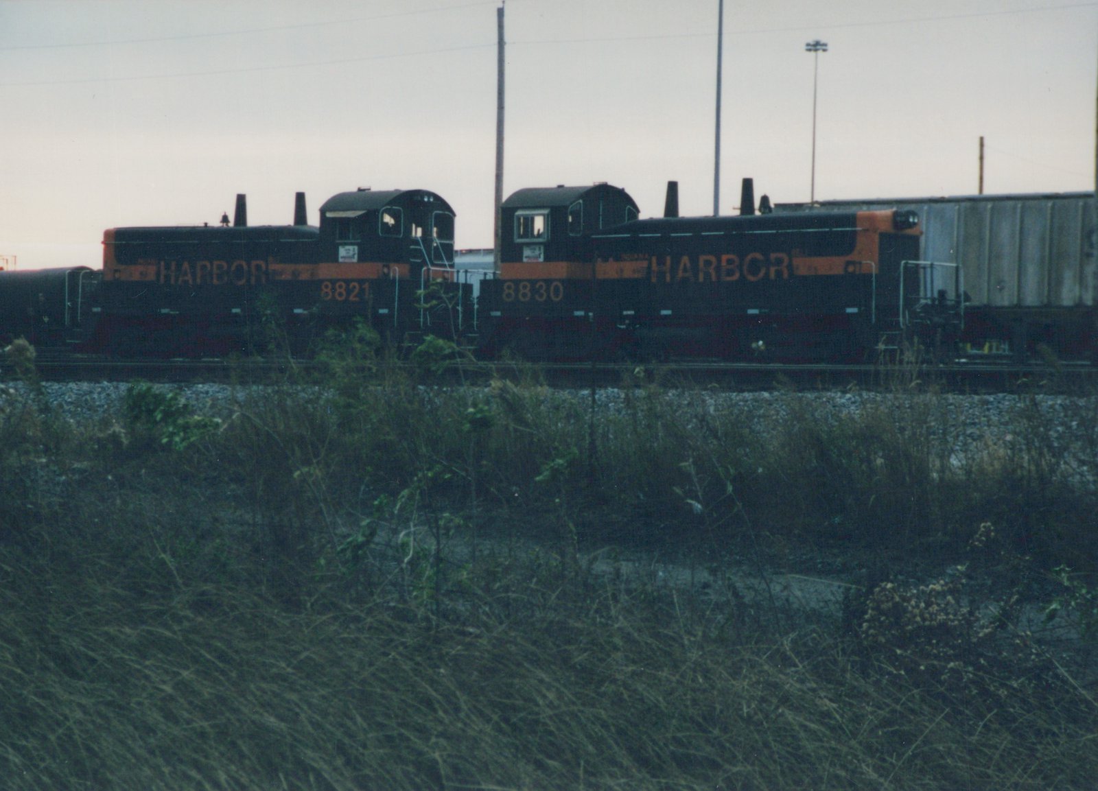 Two black and orange Indiana Harbor Belt Railroad diesel locomotives, numbered 8830 and 8821, are parked side by side in a grassy rail yard under an overcast sky.