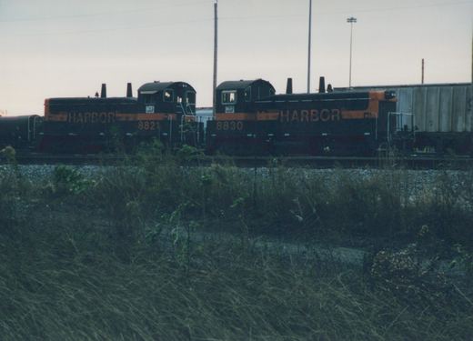 Two black and orange Indiana Harbor Belt Railroad diesel locomotives, numbered 8830 and 8821, are parked side by side in a grassy rail yard under an overcast sky.