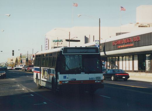 A white, red, and blue Chicago Transit Authority bus, number 5493, drives down a city street with storefronts and a black car nearby.